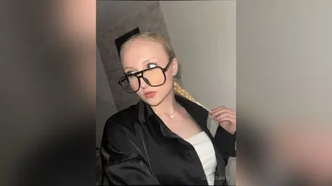 Blonde woman in black satin shirt and glasses posing with a confident expression in a modern indoor setting.