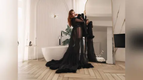 Elegant woman in a flowing black sheer gown posing in a bright, modern minimalist bathroom with a large mirror.