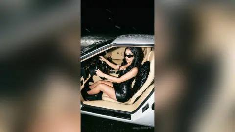 Stylish woman in a black dress and sunglasses driving a luxury car at night.