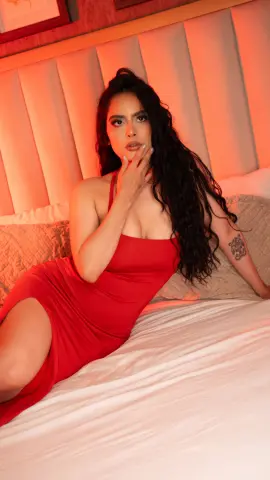 Glamorous woman in a red cocktail dress sitting on a bed with warm, moody lighting.