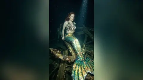 Redheaded mermaid in a shimmering gold scale tail resting on a dark underwater shipwreck ruins.