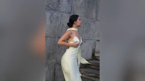 Elegant woman in a pale yellow halter neck gown standing against a rustic stone wall in soft natural light.