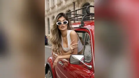 Stylish blonde woman in white tank top and sunglasses leaning out of a vintage red car on a city street.