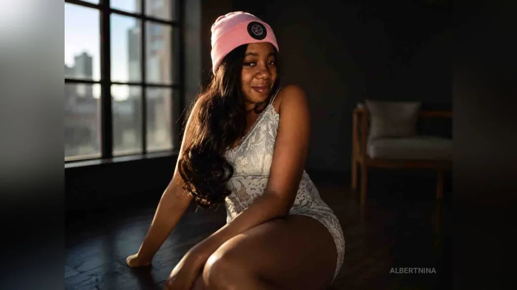 AlbertNina — AlbertNina sits on a dark floor wearing a white lace slip and a pink beanie near a window.