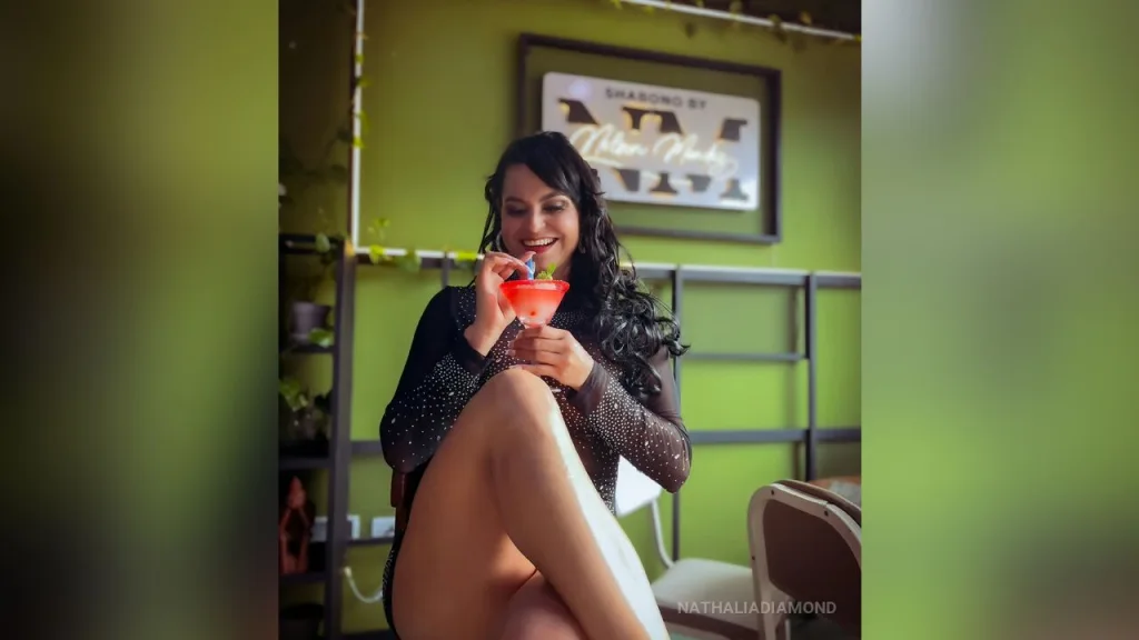 NathaliaDiamond — NathaliaDiamond sits in a black mesh bodysuit while sipping a red cocktail in a green room.