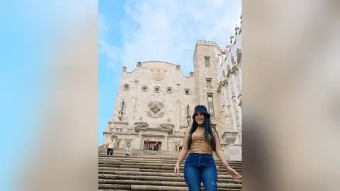 Smiling woman in a brown tank top and jeans standing before a historic stone cathedral under a blue sky.