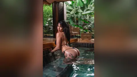 Brunette woman in black bikini relaxing in a tropical outdoor pool surrounded by lush green palm trees.