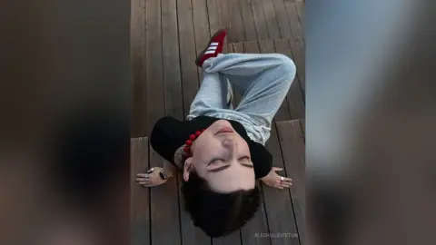 Young woman in a black t-shirt and grey sweatpants relaxing on a wooden deck in a casual, laid-back mood.