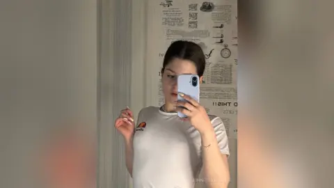 Young woman in a white t-shirt taking a mirror selfie in a modern room with newspaper wallpaper.