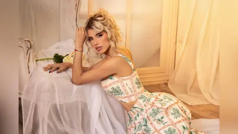 Blonde woman in a floral print crop top relaxing on a white draped chair in a bright, airy room.