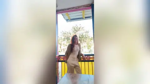 AmelieLautrec stands on a balcony with palm trees in the background, wearing a light tan tank top and matching tan trousers with an open tan cardigan.