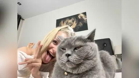 Happy blonde woman making a peace sign next to a large grey British Shorthair cat in a bright bedroom.