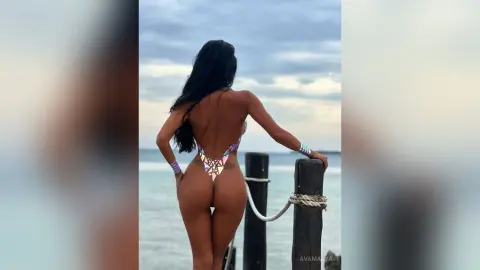 Fit woman in a colorful patterned bikini standing by a wooden pier overlooking a calm ocean at dusk.