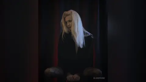 Blonde woman in a black long-sleeve top and patterned tights sitting in a moody, dark studio setting.