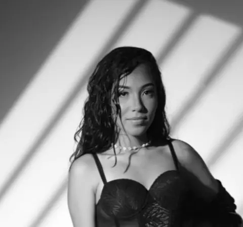 Moody black and white portrait of a woman with wavy hair wearing a lace bra in dramatic sunlight shadows.