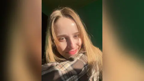 Smiling young woman with long blonde hair wearing a cozy plaid flannel shirt in warm sunlight.