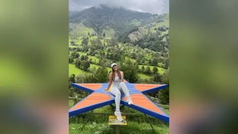 Smiling woman in a grey tank top and white leggings sitting on a large blue star sculpture in a lush green valley.