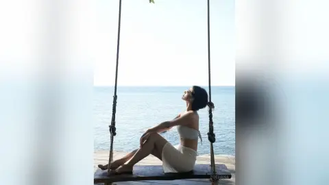Relaxed woman in a white bikini sitting on a wooden swing overlooking the calm blue ocean under bright sunlight.