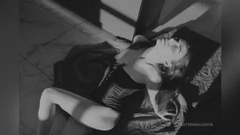 Moody black and white portrait of a woman in a dark dress lying on a textured bed in dramatic sunlight.