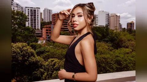 Stylish woman in a black sleeveless dress posing on a balcony overlooking a lush green city skyline.