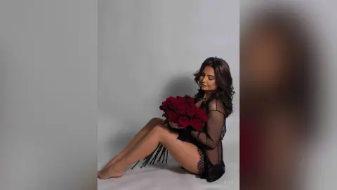 Elegant woman in a black lace outfit holding a bouquet of red roses against a white studio background.