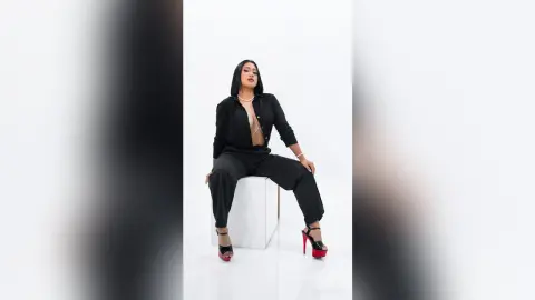 KimRoberth sits on a white cube wearing a black button-up shirt and black trousers with red high heels.