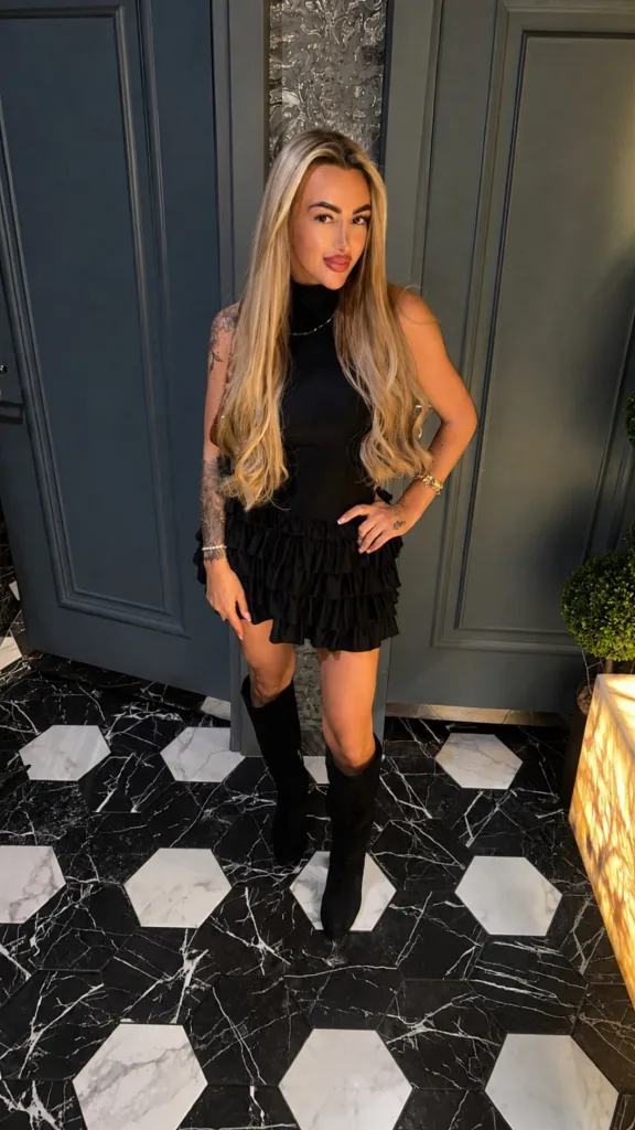 Blonde woman in a chic black dress and tall boots taking a mirror selfie in a bright bedroom.