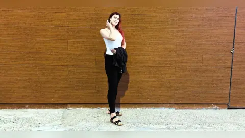 Redheaded woman in a white tank top and black leggings standing against a wooden wall background.