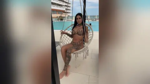 Tattooed woman in leopard print bikini relaxing on a white hanging chair by a luxury resort swimming pool.