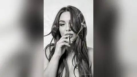 Moody black and white portrait of a young woman with long wavy hair smoking a cigarette in a studio setting.
