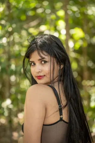 Young woman with long dark hair in a black tank top smiling in a lush green forest setting.