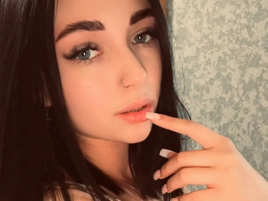 Close-up portrait of a young woman with dark hair and natural makeup posing in a cozy indoor setting.