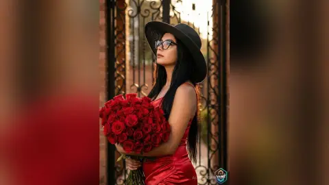 Elegant woman in a red dress holding a large bouquet of red roses in front of a vintage black iron gate.