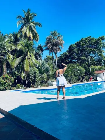 Woman in a white summer outfit standing by a tropical resort swimming pool under a bright blue sky.
