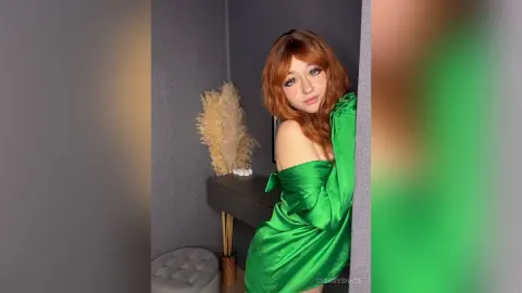 Redheaded woman in a glamorous green satin dress posing in a modern, moody interior setting.