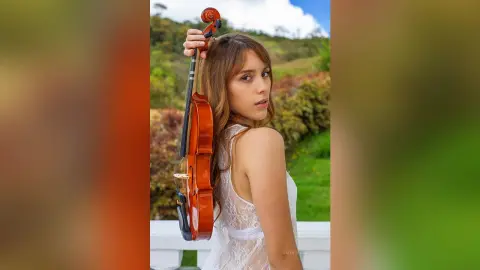 ValentinaGarcez poses outdoors in a white lace camisole, holding a wooden violin against her side.
