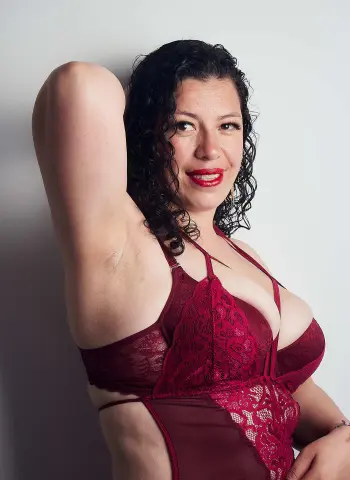 Smiling woman with curly hair wearing a red lace bodysuit against a plain white wall.