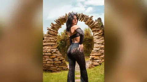 Stylish woman in a black lace cutout bodysuit and flared pants posing by a rustic stone arch in a sunny garden.