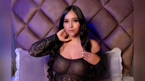 Young woman in a black lace long-sleeve top posing against a tufted headboard in soft moody lighting.