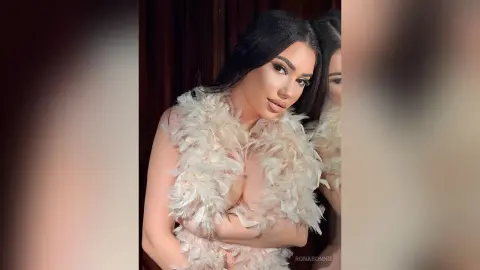 Glamorous woman in a luxury cream feathered outfit posing in a moody, elegant indoor setting.