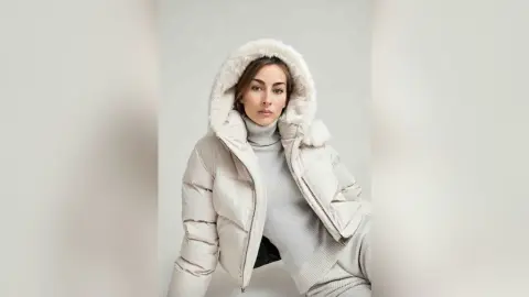 Young woman in a cozy white puffer jacket and grey turtleneck sweater posing against a neutral studio background.