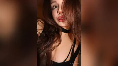 Young woman with long wavy hair wearing a black choker and top in a moody, dimly lit indoor setting.