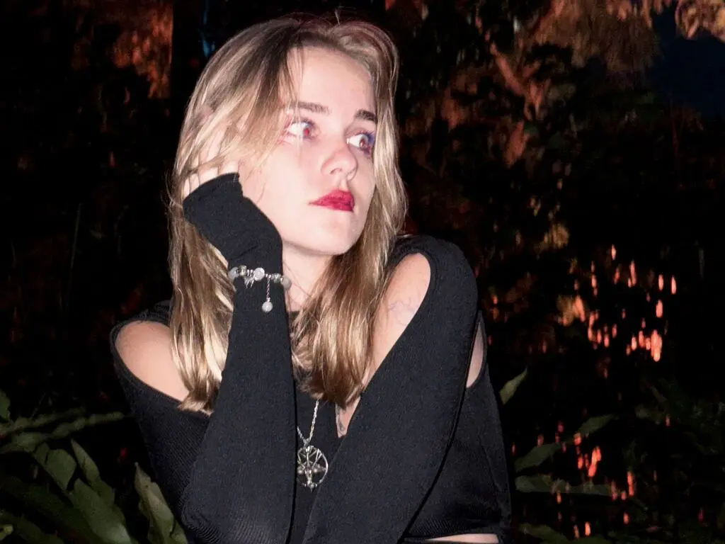 Young woman with blonde hair in a black long-sleeve top posing outdoors at night in a moody garden setting.