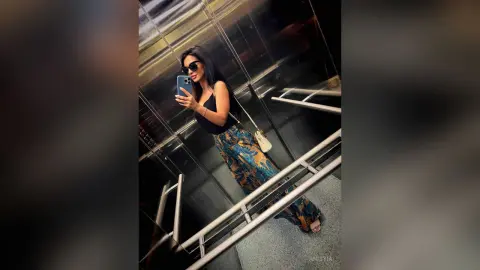 Stylish woman in black tank top and patterned trousers taking a mirror selfie in a modern elevator.