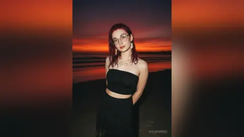 Young woman in a black strapless top and glasses at a moody beach during a dramatic orange sunset.