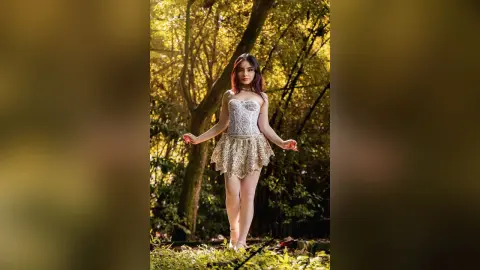 Elegant woman in a white lace corset and gold skirt standing in a sunlit, dreamy forest setting.
