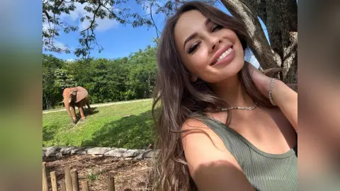 EvelynMills smiles while leaning her head back against a large tree trunk in an outdoor grassy setting.