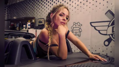 Blonde woman in black lingerie leaning on a car in a modern studio with a playful mood.