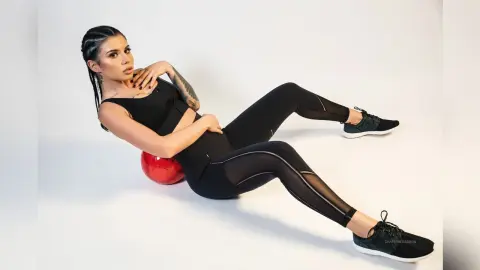 Athletic woman in black yoga outfit and sneakers sitting on a red gym ball in a bright studio setting.