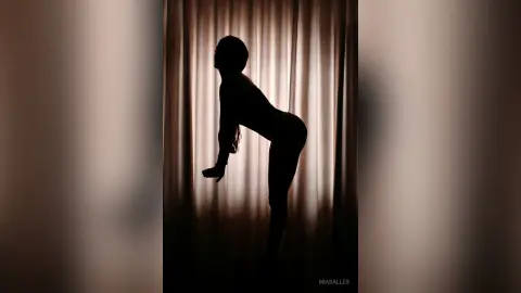 Silhouette of a woman in a bodysuit posing against backlit curtains in a moody, minimalist indoor setting.